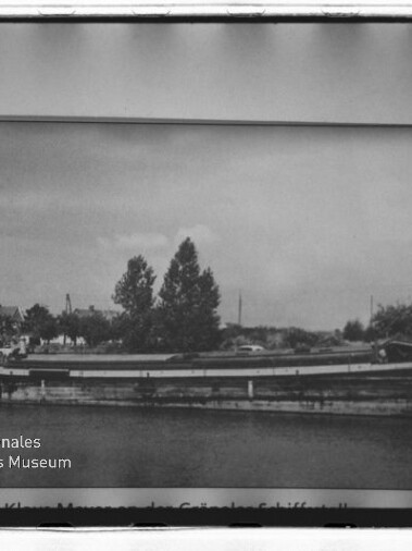 Schwarzweiß-Foto eines langen Binnenschiffs am Ufer vor Bäumen und Häusern, gerahmt; Schriftzug Internationales Maritimes Museum