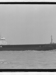 Schwarzweiß-Seitenansicht eines Frachtschiffs mit Aufbauten achtern auf ruhiger See, Museumslogo links unten