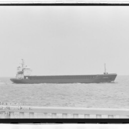 Langgestrecktes Frachtschiff in Seitenansicht auf ruhigem Meer; Reling im Vordergrund und Museumswasserzeichen links unten.