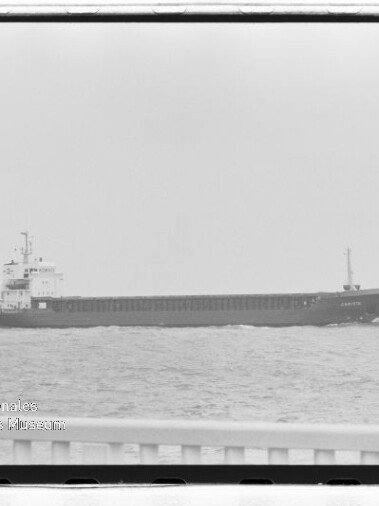 Langgestrecktes Frachtschiff in Seitenansicht auf ruhigem Meer; Reling im Vordergrund und Museumswasserzeichen links unten.