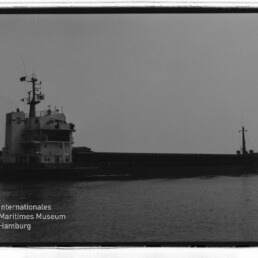 Silhouette eines Frachtschiffs mit achterlichem Aufbau auf ruhigem Wasser, Museumslogo unten links.