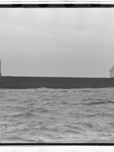 Langes Frachtschiff auf bewegter See unter grauem Himmel, mit Internationales Maritimes Museum Hamburg‑Wasserzeichen unten links.