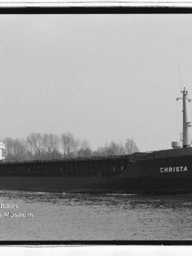 Frachtschiff CHRISTA auf einem Kanal, seitlich zu sehen, dunkler Rumpf, weiße Aufbauten und Baumreihe im Hintergrund.