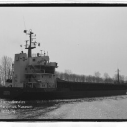 Frachtschiff mit hohen Aufbauten und Funkmast, Kennzeichnung J-3, fährt auf ruhigem Wasser vor kahlen Uferbäumen.