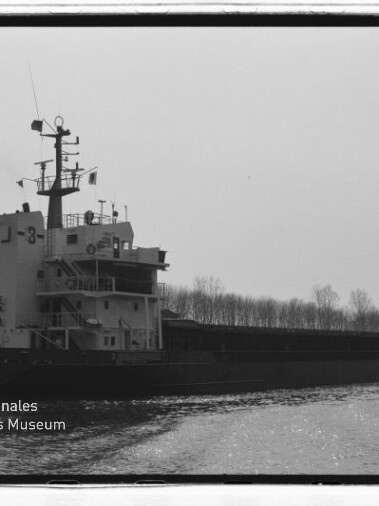 Frachtschiff mit hohen Aufbauten und Funkmast, Kennzeichnung J-3, fährt auf ruhigem Wasser vor kahlen Uferbäumen.