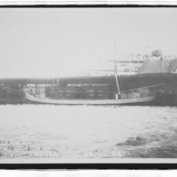 Längliches Segelboot mit zwei Masten liegt längsseits an einem Kai vor Industriegebäuden; Wasser im Vordergrund, Schwarzweiß.