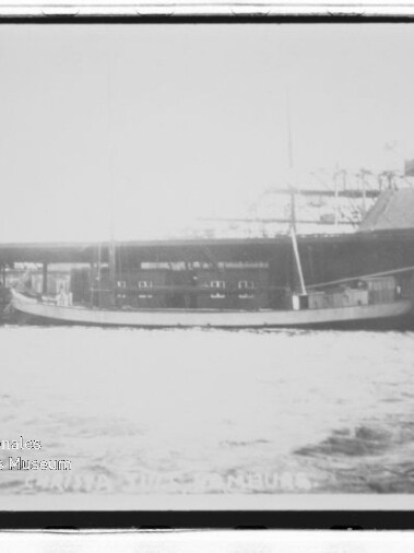 Längliches Segelboot mit zwei Masten liegt längsseits an einem Kai vor Industriegebäuden; Wasser im Vordergrund, Schwarzweiß.