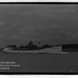 Weißes Frachtschiff mit lesbarem Namen CHRISTA 383 fährt vor Küstenlinie unter dunkler Wolkendecke; Museumslogo unten links.