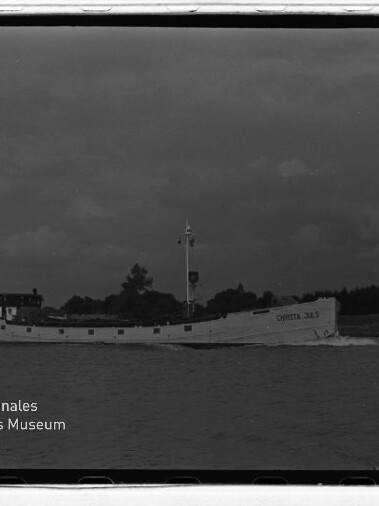 Weißes Frachtschiff mit lesbarem Namen CHRISTA 383 fährt vor Küstenlinie unter dunkler Wolkendecke; Museumslogo unten links.