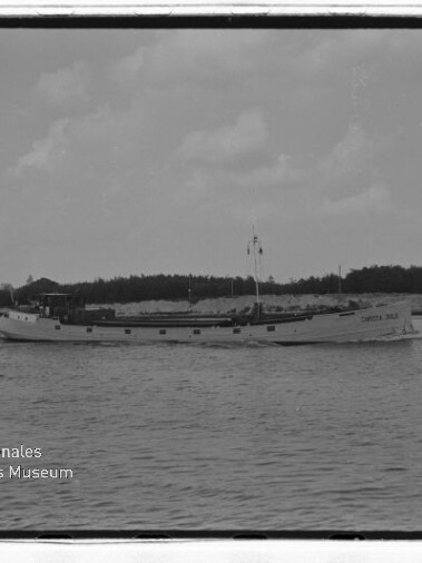 Schwarzweißfoto eines langen Küstenmotorschiffs auf ruhiger See vor baumbestandener Küste; Wasserzeichen 