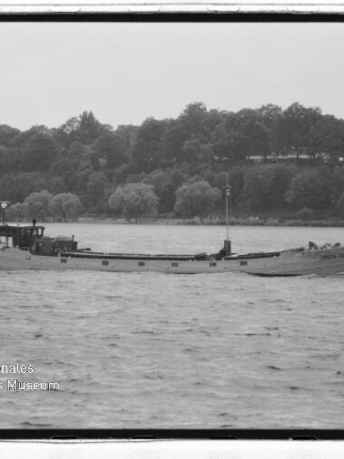Schlankes Frachtschiff fährt auf einem Fluss vor baumbewachsenem Ufer; Wasserzeichen Internationales Maritimes Museum Hamburg.