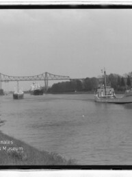 Kanal mit mehreren Frachtschiffen, ein Schiff fährt rechts, im Hintergrund eine Stahlfachwerkbrücke und Uferbäume.