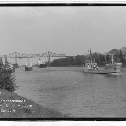Kanal mit mehreren Frachtschiffen, ein Schiff fährt rechts, im Hintergrund eine Stahlfachwerkbrücke und Uferbäume.