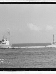 Schwarzweißaufnahme eines langen Frachtschiffs mit erhöhtem Achteraufbau auf ruhiger See, Museumswasserzeichen unten links.