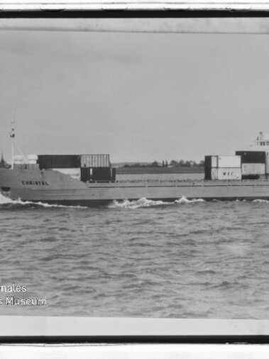 Frachtschiff Christel mit wenigen Containern fährt über ruhige See, Küste im Hintergrund.