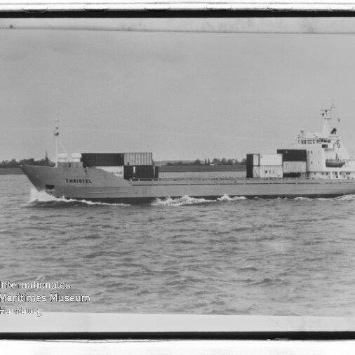 Frachtschiff Christel mit wenigen Containern fährt über ruhige See, Küste im Hintergrund.
