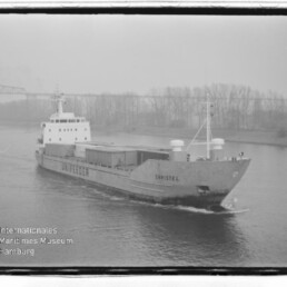 Frachtschiff Christel mit Unifeeder-Schriftzug fährt einen Fluss entlang; Brücke und Uferbäume im Nebel.