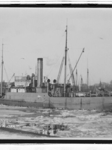 Historisches Dampfschiff mit hohem Schornstein und Kränen liegt in eisbedecktem Hafenbecken, Museumslogo links unten.
