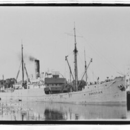 Frachtschiff Christian mit rauchendem Schornstein und Ladegeschirr am Kai, Spiegelung im ruhigen Hafenwasser.