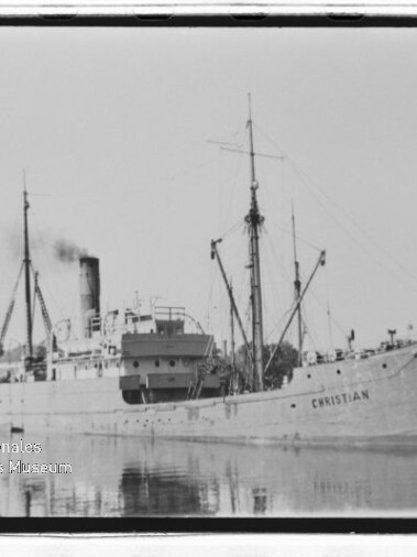 Frachtschiff Christian mit rauchendem Schornstein und Ladegeschirr am Kai, Spiegelung im ruhigen Hafenwasser.