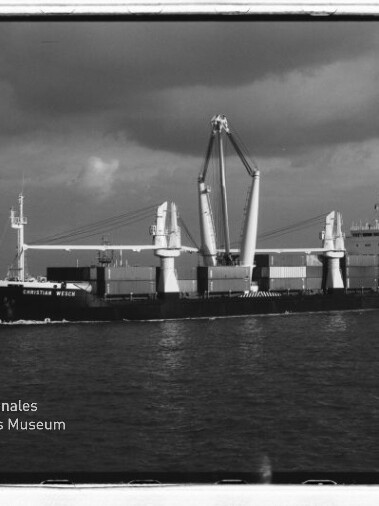 Schwarzweißaufnahme eines Frachtschiffs mit zwei großen Hubkränen und Containerstapeln auf ruhiger See; Museumslogo unten links.