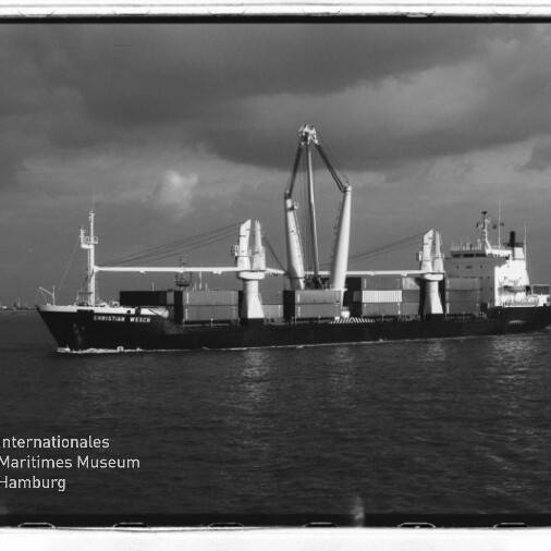 Schwarzweißaufnahme eines Frachtschiffs mit zwei großen Hubkränen und Containerstapeln auf ruhiger See; Museumslogo unten links.