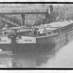 Schwarzweißaufnahme eines Lastkahns mit den Namen CHRISTINA und WILSTER, längsseits an einem hölzernen Anlegesteg