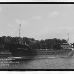 Schwarzweißfoto eines Frachtschiffs namens CHRISTINA 805, das vor einem baumbewachsenen Ufer fährt; Museumslogo unten links.