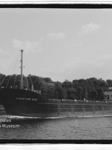 Schwarzweißfoto eines Frachtschiffs namens CHRISTINA 805, das vor einem baumbewachsenen Ufer fährt; Museumslogo unten links.