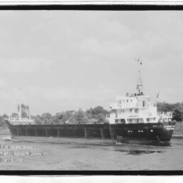 Schwarzweißfoto eines Frachtschiffs auf einem Kanal mit bewaldetem Ufer; Schriftzug Internationales Maritimes Museum Hamburg links unten.