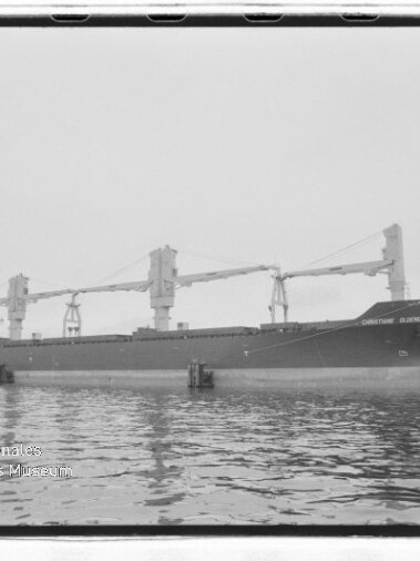 Großes Frachtschiff mit drei Ladekränen liegt im Hafen, ruhige Wasseroberfläche und Museumslogo unten links.