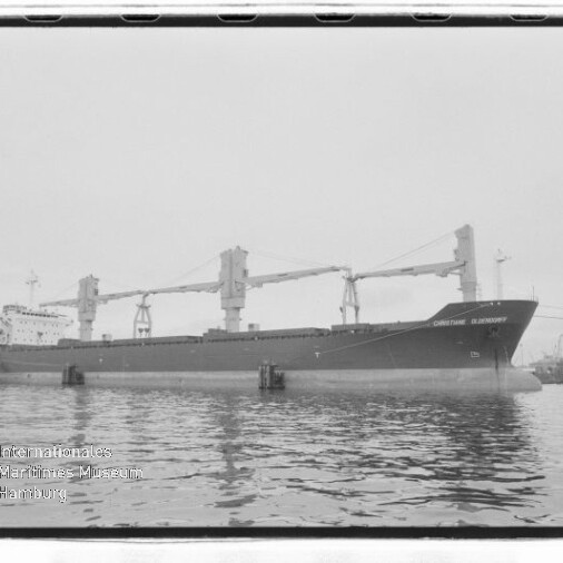 Großes Frachtschiff mit drei Ladekränen liegt im Hafen, ruhige Wasseroberfläche und Museumslogo unten links.
