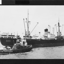 Schwarzweißaufnahme eines Frachtschiffs namens ALGOL HAMBURG mit Ladekränen, begleitet von einem Schlepper im Hafen.