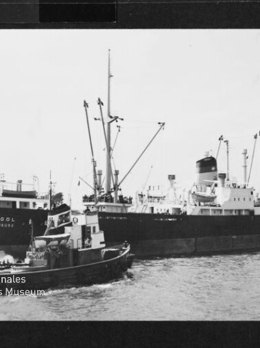 Schwarzweißaufnahme eines Frachtschiffs namens ALGOL HAMBURG mit Ladekränen, begleitet von einem Schlepper im Hafen.