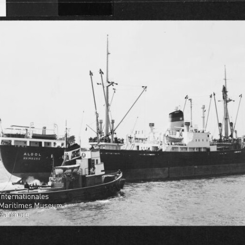 Schwarzweißaufnahme eines Frachtschiffs namens ALGOL HAMBURG mit Ladekränen, begleitet von einem Schlepper im Hafen.