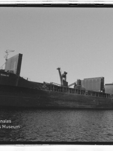 Frachtschiff längsseits an einem Industriehafen mit Kränen und Förderanlagen, Schiffsname am Bug sichtbar.