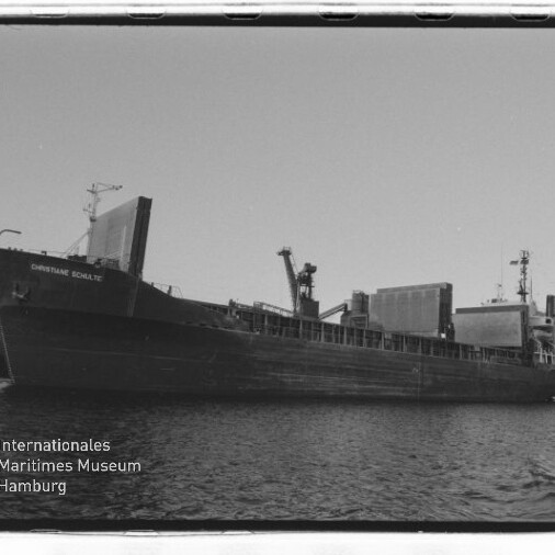 Frachtschiff längsseits an einem Industriehafen mit Kränen und Förderanlagen, Schiffsname am Bug sichtbar.