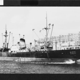 Schwarzes Frachtschiff mit Ladekränen erzeugt Bugwelle, Hafen- und Lagerhallen im Hintergrund.