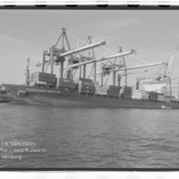 Containerschiff CHRISTIANE SCHULTE am Kai unter großen Hafenkränen, Containerstapeln und Wasser vorn; Museumslogo sichtbar.