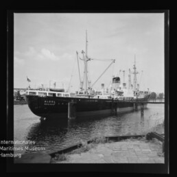 Großes Frachtschiff mit Masten und Ladekränen längsseits an einem Kai, Wasserfläche und Museumssiegel in der Ecke.
