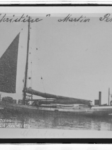 Ein kleines Einmast-Segelschiff mit aufgeholtem dreieckigem Segel liegt ruhig im Wasser, oben ist ein handschriftlicher Schriftzug sichtbar.