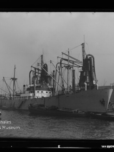 Schwarzweißfoto eines großen Frachtschiffs mit hohen Schornsteinen und Kränen im Hafen, kleine Begleitboote am Bug.