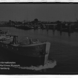 Frachtschiff im Hafen mit Bug im Vordergrund, Industrie am Ufer und Wasserzeichen 'Internationales Maritimes Museum Hamburg'.