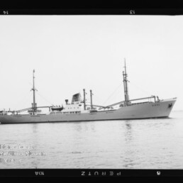 Frachtschiff mit zwei Masten, mittigem Schornstein und Ladekränen auf ruhiger See, schwarzweiß fotografiert.