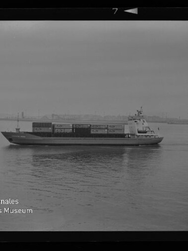 Containerschiff in Seitenansicht auf ruhiger See mit gestapelten Containern und schwacher Hafen-Silhouette am Horizont.