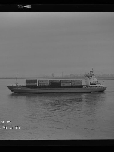 Containerschiff fährt auf ruhigem Wasser, Containerstapel auf dem Deck und undeutliche Hafenkulisse im Hintergrund.