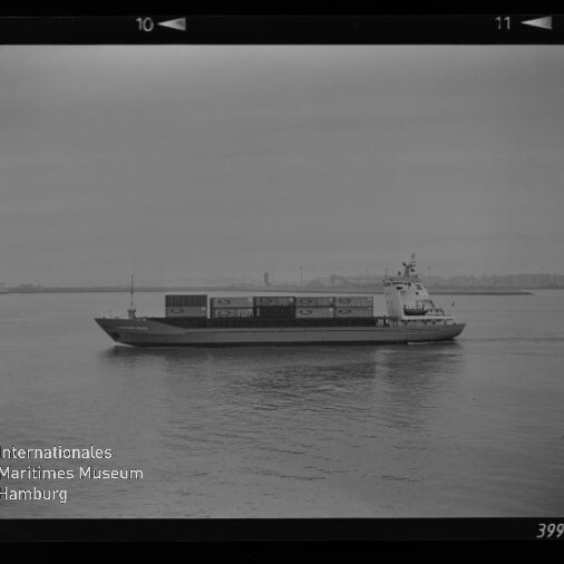 Containerschiff fährt auf ruhigem Wasser, Containerstapel auf dem Deck und undeutliche Hafenkulisse im Hintergrund.