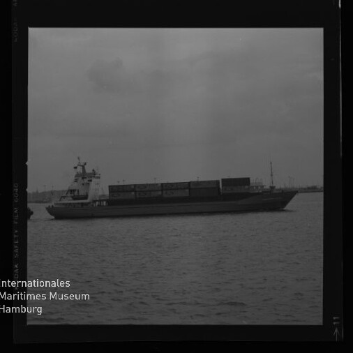 Containerschiff seitlich auf ruhigem Wasser vor Hafen bei bewölktem Himmel, Museum-Logo unten links