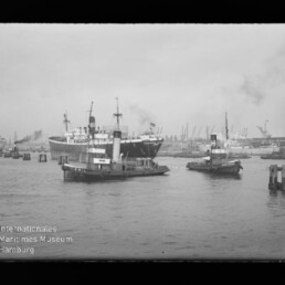 Zwei Schlepper im Vordergrund, großes Passagierschiff im Hafen mit Hafenkränen im Hintergrund; Wasserzeichen Internationales Maritimes Museum Hamburg.