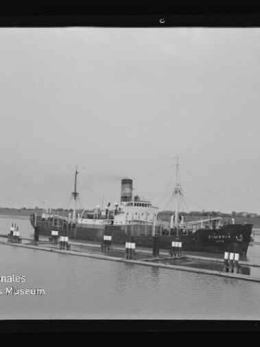 Dampffrachter mit Schornstein und Ladegeschirr am Pier in einer Schwarzweißaufnahme, Schriftzug Internationales Maritimes Museum Hamburg sichtbar